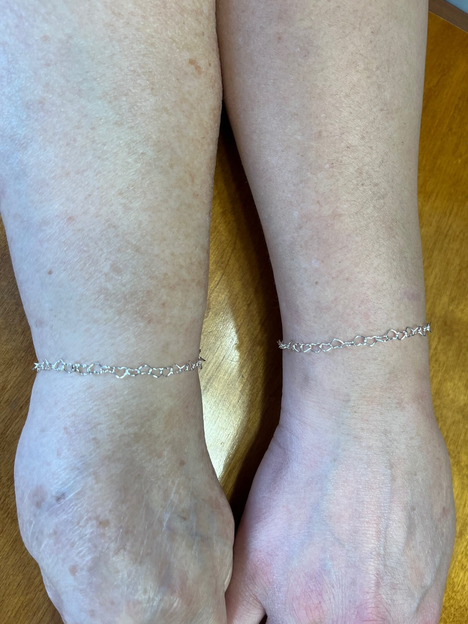 Two wrists wearing matching silver heart-link bracelets
